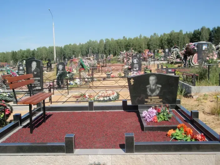 Памятник арочной формы из чёрного гранита на площадке с декоративной красной каменной крошкой и скамьёй